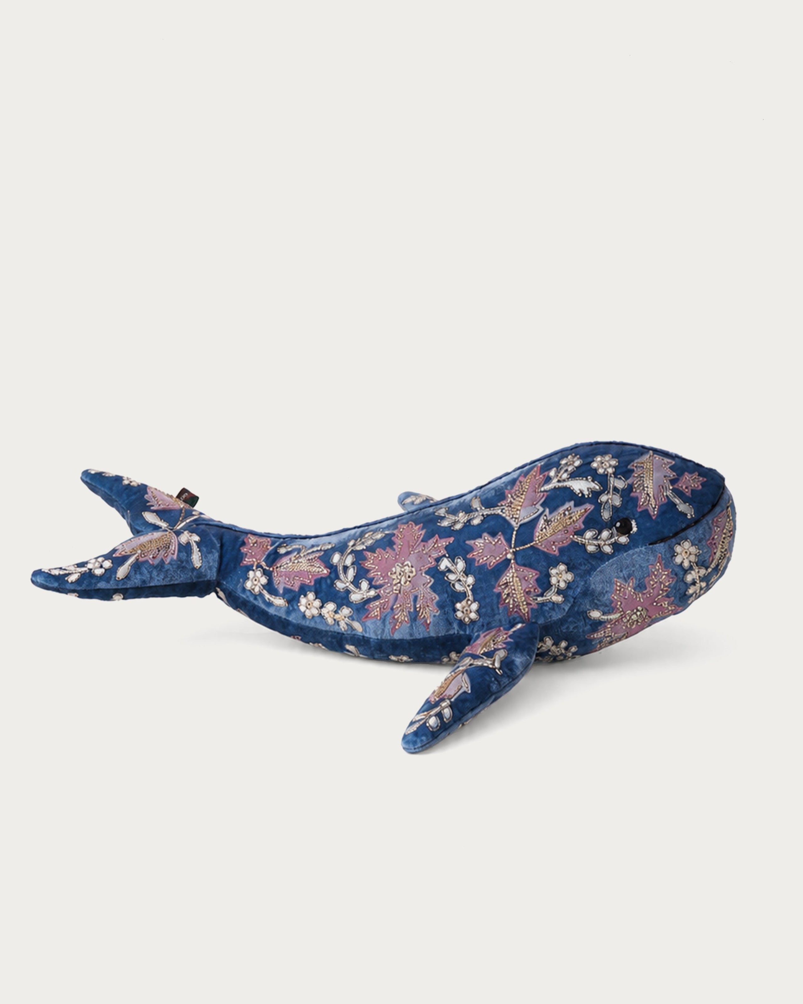 A whale-shaped plush toy or pillow featuring a blue floral pattern with pink and white blossoms, set against a plain light gray background.