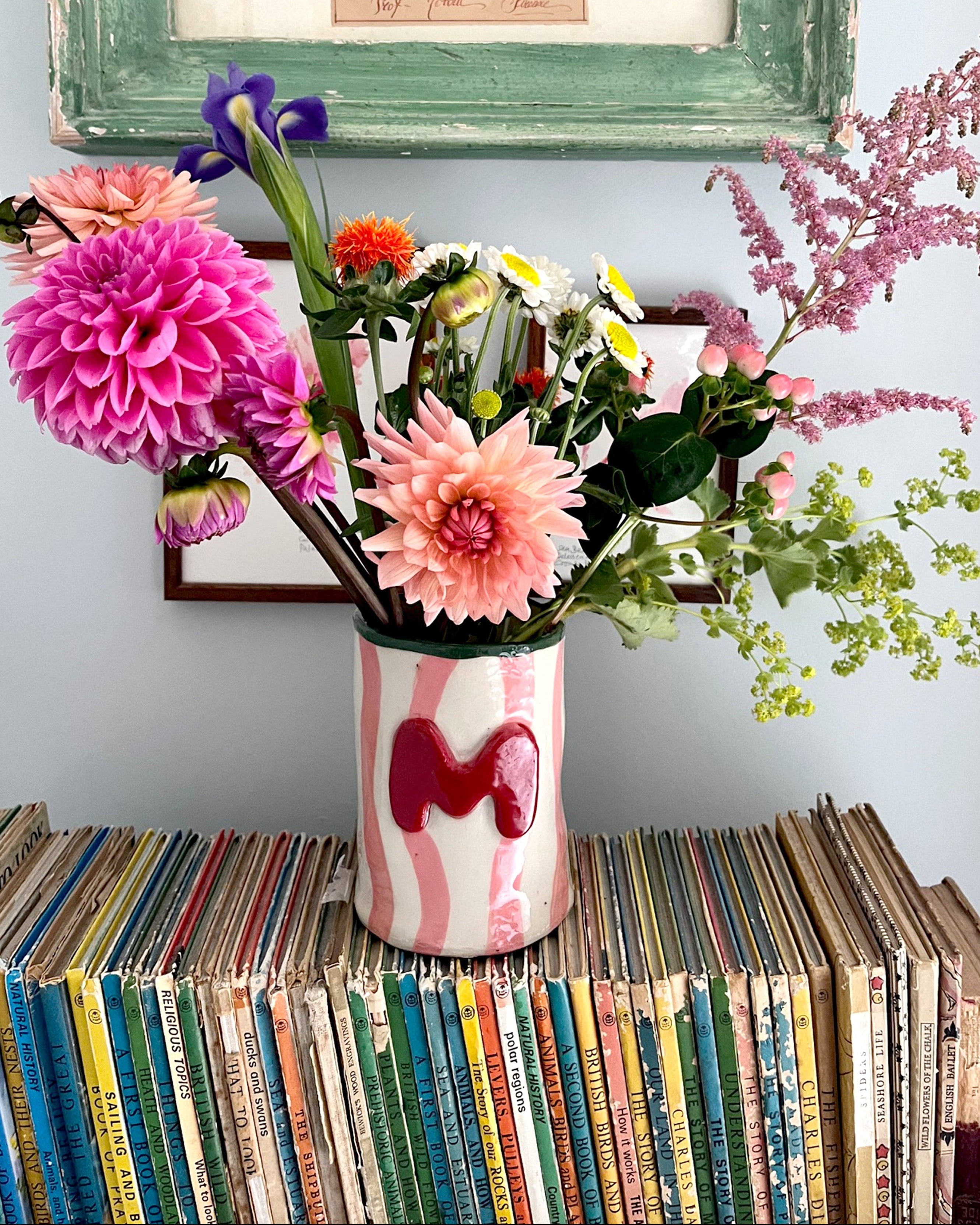 A ceramic vase with pink and white wavy stripes and a red heart, filled with a colorful bouquet of flowers, sitting on top of a stack of vintage record sleeves against a light blue wall.
