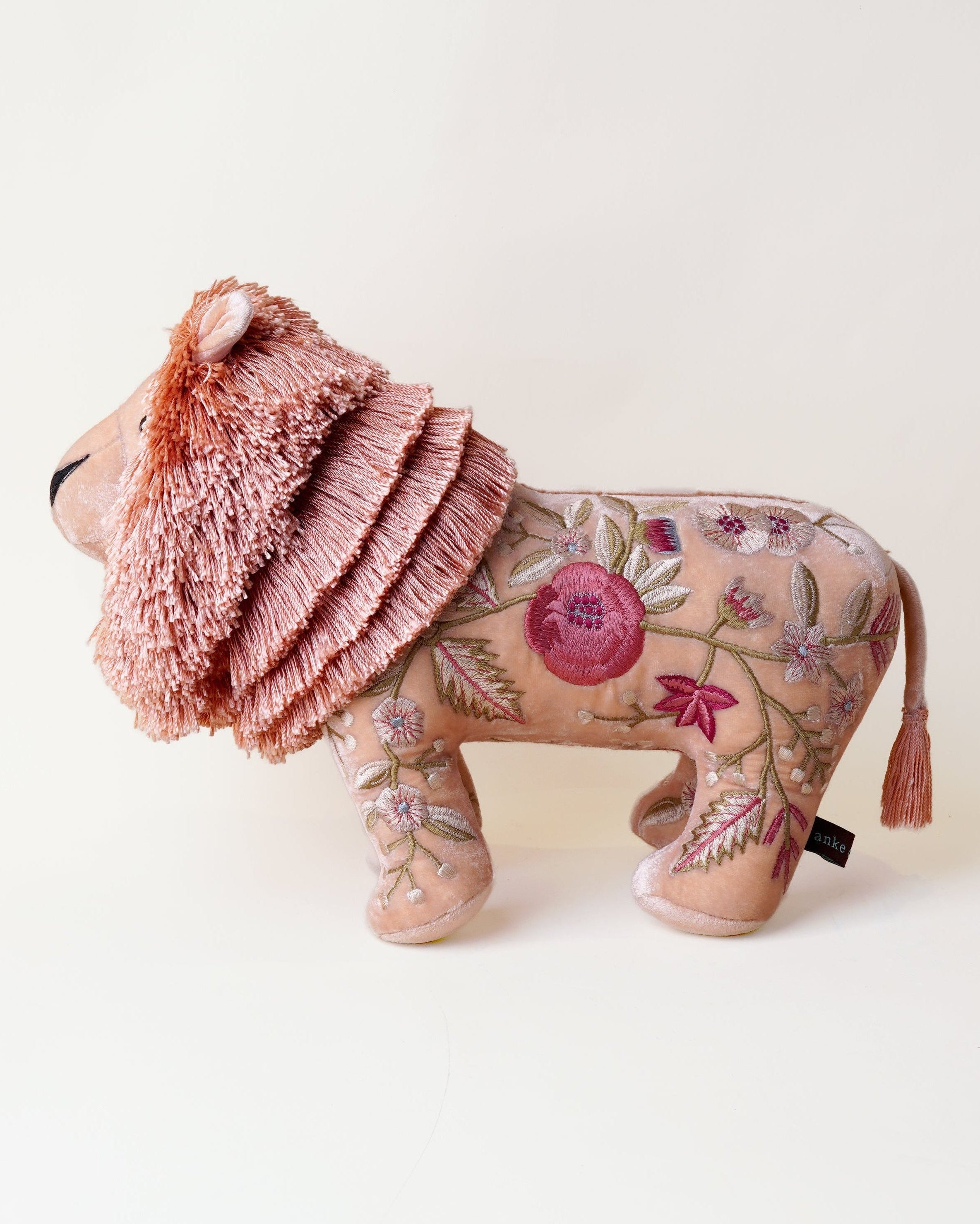 A decorative lion-shaped plush toy or cushion in dusty pink, featuring a textured pink mane, embroidered floral pattern on its body, and a small tassel on the tail, photographed against a plain off-white background.
