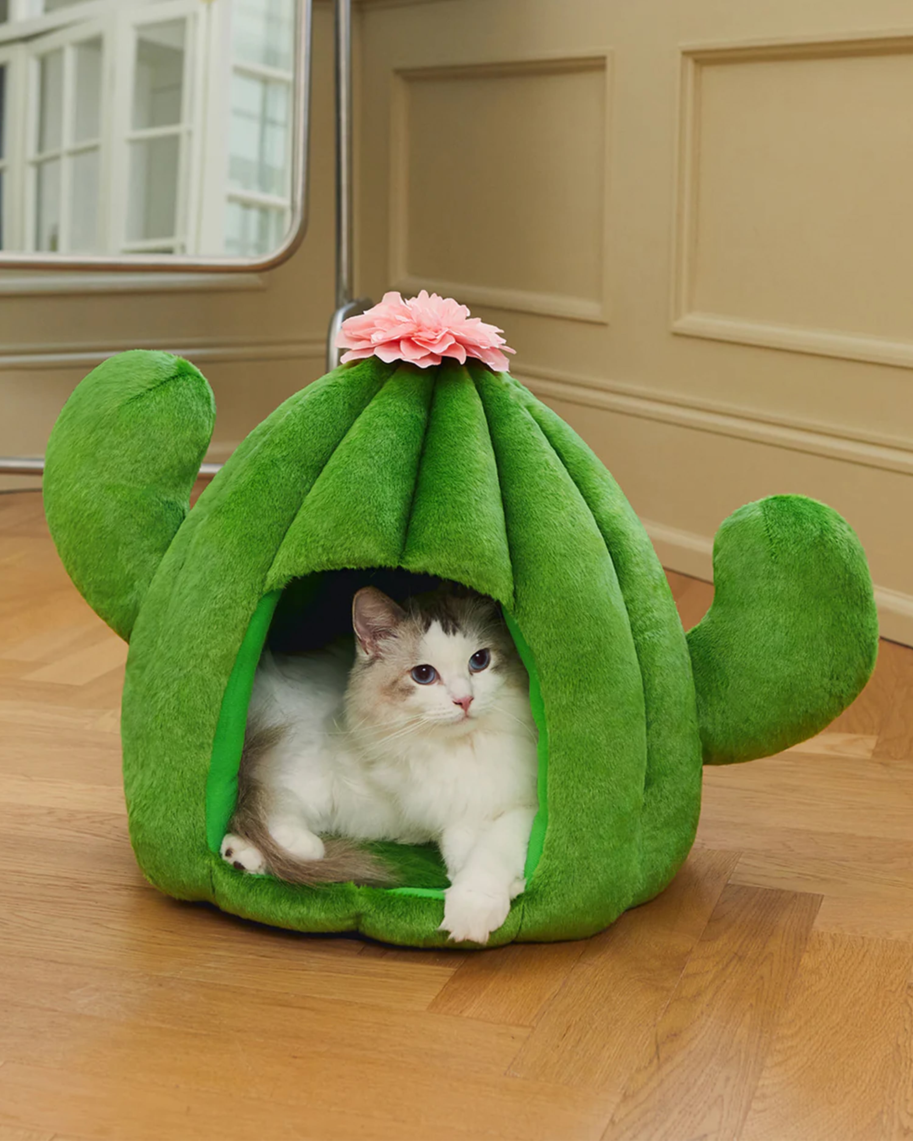 Cat inside a green cactus-shaped pet bed with a pink flower on a wooden floor.