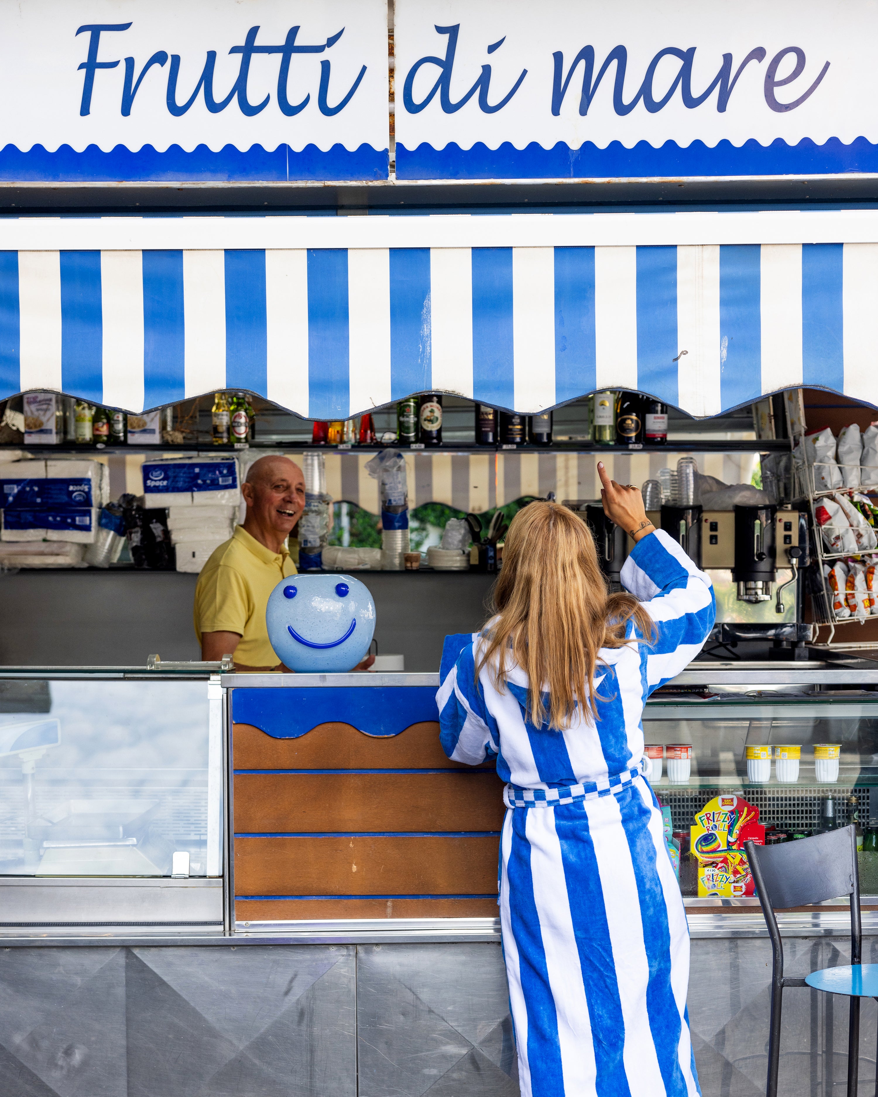 Woman in a blue and white striped dress interacting with a man behind a counter with a smiley face vase on the counter, under a blue and white striped awning with 'Frutti di mare' text.