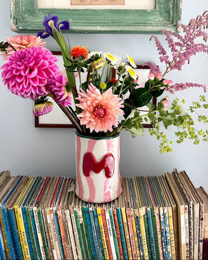 A ceramic vase with pink and white wavy stripes and a red heart, filled with a colorful bouquet of flowers, sitting on top of a stack of vintage record sleeves against a light blue wall.