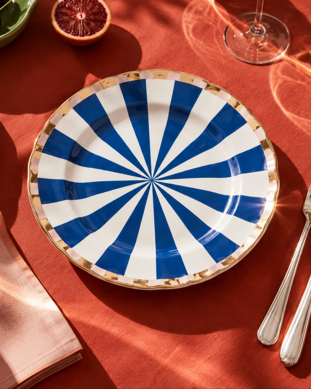 A round ceramic plate with a blue and white radial striped pattern and a gold rim, styled on a red tablecloth with silver cutlery, a wine glass, and fruit.