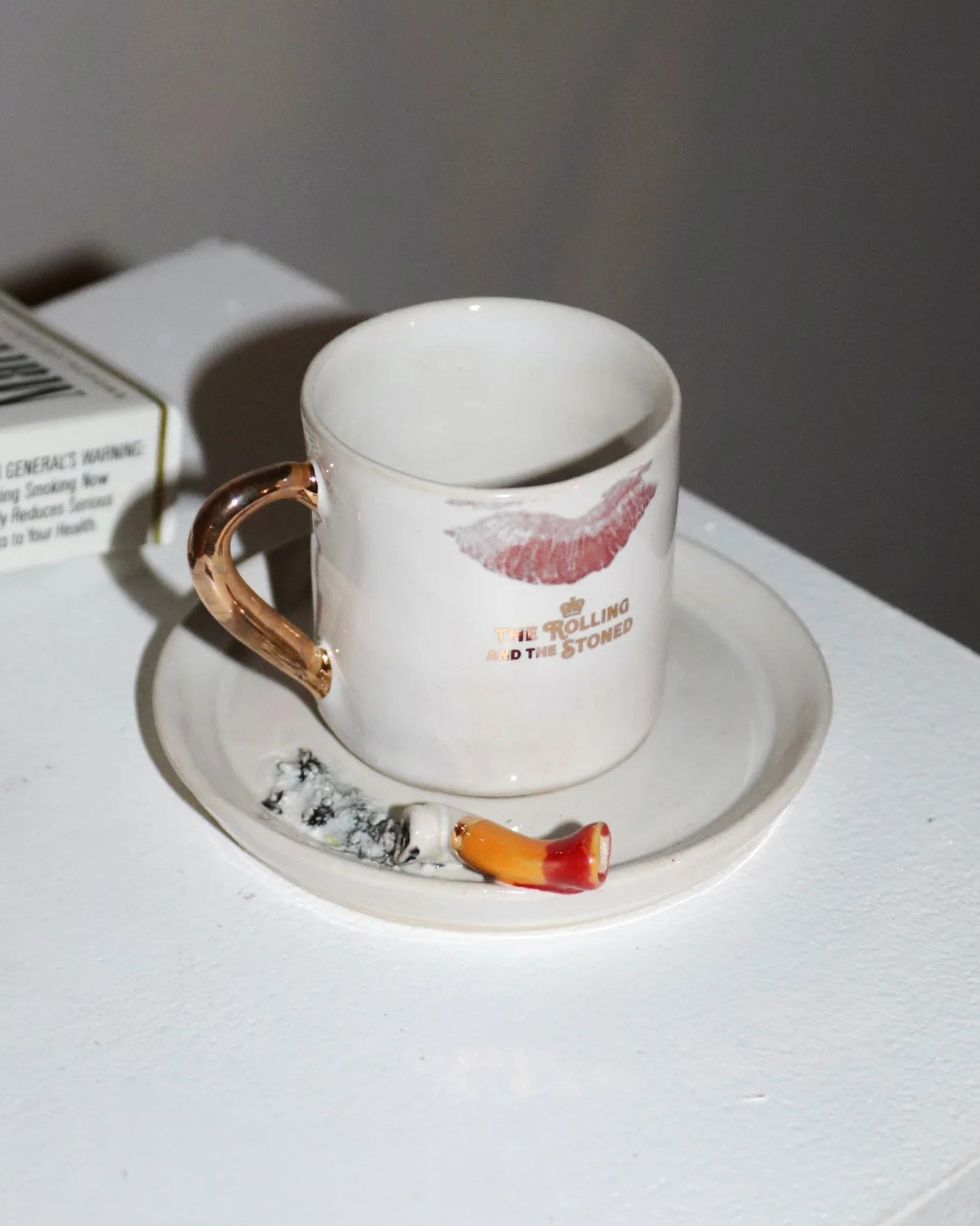 White mug with red lip print and gold handle on a matching saucer with cigarette butt, against a white background.