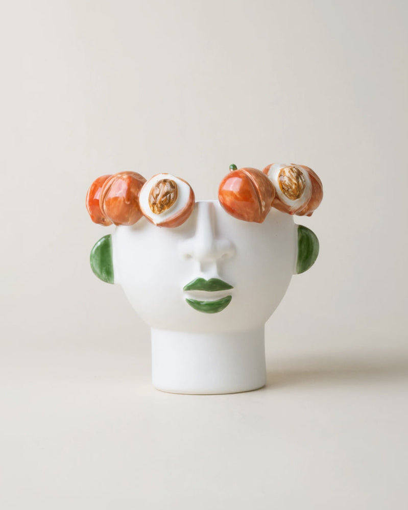 A small white vase in the shape of a stylized face with peaches arranged around the crown of the vase, resembling eyes and a mouth.