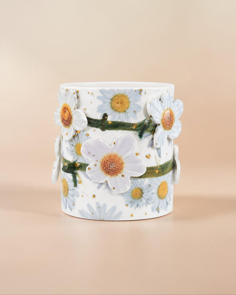 A white cylindrical ceramic vase decorated with 3D white daisies, blue daisies, green stems, and tiny gold speckles, set against a solid beige background.