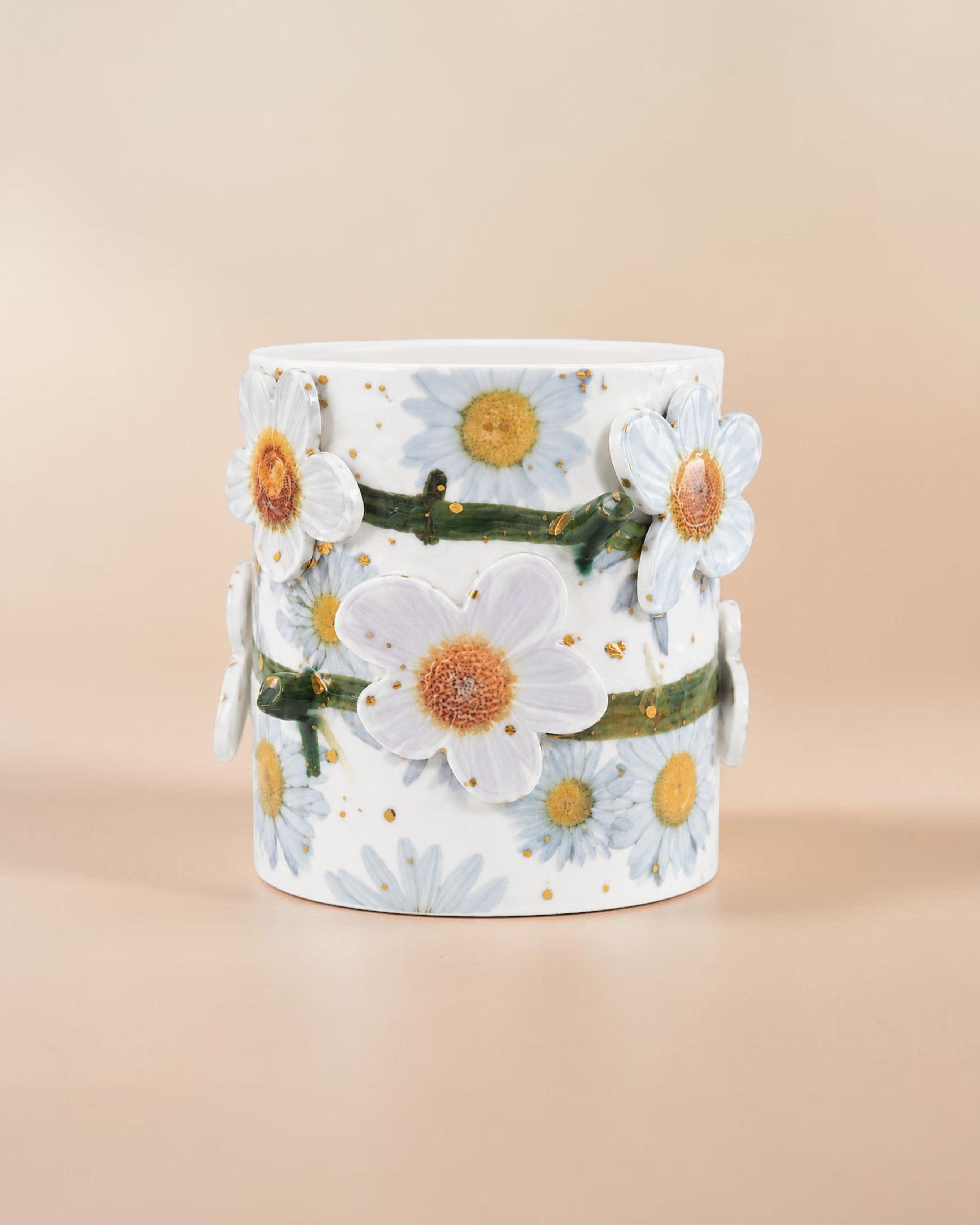 A white cylindrical ceramic vase decorated with 3D white daisies, blue daisies, green stems, and tiny gold speckles, set against a solid beige background.